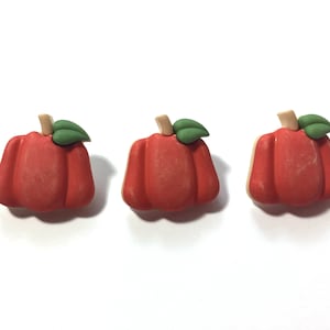 May include: Three small, red pumpkin-shaped decorative objects with green leaves and tan stems. The pumpkins are arranged in a row against a white background, perfect for fall or Halloween decor.