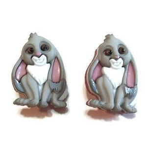 May include: A pair of gray bunny-shaped earrings. Each earring features a sitting bunny with large ears, pink inner ears, and a white chest. The bunnies have brown eyes and are set against a white background.