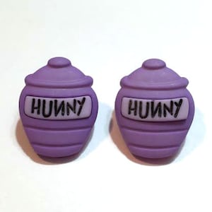 May include: Two purple pot-shaped charms with lids, each bearing the word "HUNNY" in black letters on a white background. The charms are a light purple color and have horizontal lines around the body of the pot.