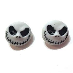 May include: A pair of white stud earrings featuring the face of Jack Skellington from The Nightmare Before Christmas. Each earring has black eyes and a stitched smile.