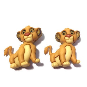 May include: Two Simba-shaped buttons. Each button features a light brown lion cub with a cream-colored chest, a red nose, and large, expressive eyes. The buttons are likely for sewing or crafting projects.