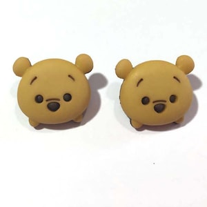 May include: Two Winnie the Pooh-themed charms. Each charm is a light brown color with black eyes and a black nose. The charms have small, rounded ears and are set against a white background. These charms are likely for crafting or jewelry making.