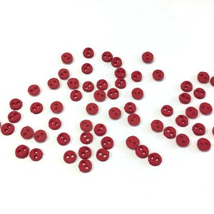 May include: A collection of small, round, red buttons with two holes each, scattered on a white surface. The buttons are evenly colored and appear to be made of wood or a similar material. Ideal for sewing and craft projects.