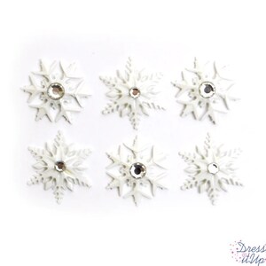 May include: Six white snowflake embellishments with a central clear rhinestone. The snowflakes have detailed, delicate designs and are arranged on a white background. The image includes the text "Dress It Up".
