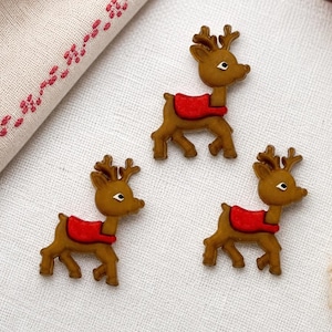May include: Three brown reindeer-shaped buttons with red saddles. Each reindeer has a light brown body, dark brown antlers, and a bright red saddle across its back. The buttons are arranged on a white fabric surface near a spool of red thread.