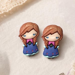 May include: A pair of stud earrings shaped like small figures with brown braided hair, blue eyes, and wearing a blue dress with a pink bodice and light blue trim. The figures have a smooth, matte finish and are crafted with detailed facial features.