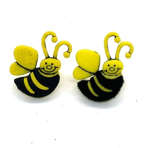 May include: Two bee-shaped earrings. Each earring features a yellow head and wings, with a black and yellow striped body. The bees have curved antennae and smiling faces. The earrings are set against a white background.