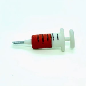 May include: A white and red syringe with a silver needle. The syringe has a red plunger with black lines and a white body. The needle is silver and pointed. The syringe is lying on a white surface.