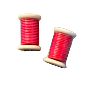 May include: Two cylindrical spools of bright pink thread, each featuring white plastic ends. The spools are positioned at slight angles, showcasing the tightly wound, vibrant pink thread wrapped around the central core of each individual spool.