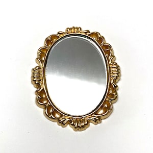 May include: A small, oval-shaped mirror with a gold-colored ornate frame. The frame features a detailed, decorative design. The mirror reflects light, and the background is white. The item is likely a decorative accessory.