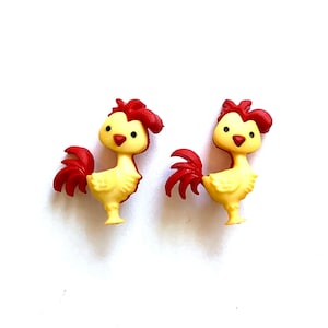 May include: Two yellow rooster-shaped embellishments with red combs and tail feathers. The rooster figures have black eyes and are standing upright. The background is white.