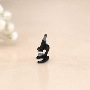 May include: A miniature black microscope with a silver eyepiece and a silver stage. The small scientific instrument features a curved base, a central vertical column, and a rotating nosepiece, rendered in a solid black finish with metallic silver accents.
