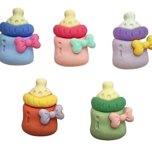 May include: A collection of five colorful baby bottle-shaped embellishments. Each bottle features a different color scheme, including blue, pink, purple, orange, and green, with matching bow accents. The tops are yellow.