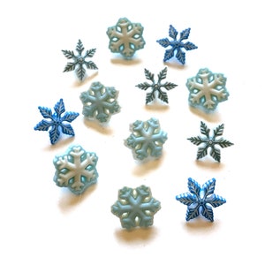 May include: A collection of snowflake-shaped embellishments in various shades of blue and white. The snowflakes are scattered across a white background, showcasing their intricate designs. These decorative elements are ideal for winter-themed crafts and projects.