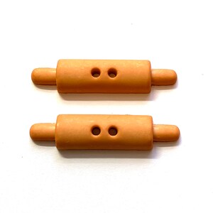 May include: Two light brown rolling pin shaped buttons with two holes each. The buttons have rounded ends and are set against a white background. These buttons are ideal for sewing and craft projects.