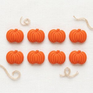May include: Eight small, bright orange pumpkin-shaped buttons arranged in two rows of four. Each button features vertical ridges and a rounded, symmetrical form. Two pieces of thin, light tan twine are curled near the corners of the arrangement.