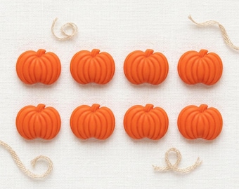 May include: Eight small, bright orange pumpkin-shaped buttons arranged in two rows of four. Each button features vertical ridges and a rounded, symmetrical form. Two pieces of thin, light tan twine are curled near the corners of the arrangement.
