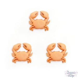 May include: Three orange crab-shaped embellishments. Each crab has a rounded body, two large claws, and small legs. The embellishments are made of a smooth material and are set against a white background. The embellishments are from Dress It Up.