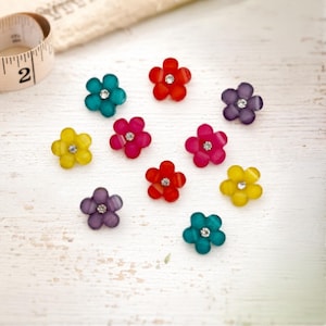 May include: A collection of ten small, five-petaled flower-shaped buttons in various colors including red, yellow, purple, pink, and teal. Each button features a small, clear, circular rhinestone set in the center. A measuring tape is visible.