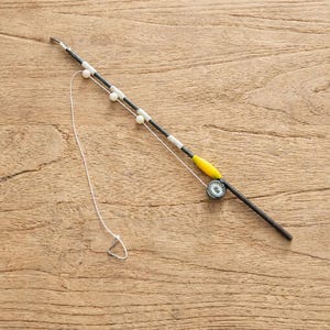 May include: A black fishing rod toy featuring a yellow float, a small silver reel, and a white string with a triangular hook. The rod is decorated with three small beads in light pink, white, and cream colors along the upper section of the shaft.