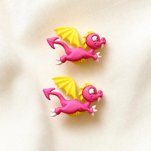 May include: Two identical pink dragon-shaped buttons featuring bright yellow wings, a yellow belly, white claws, and a single white tooth. Each dragon is depicted in a flying pose with a pointed tail, large eyes, and a small crest on its head.