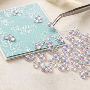 May include: A square light blue card with white embossed leaf patterns and the text Thinking of You, accompanied by a pile of iridescent, round, dome-shaped embellishments and a pair of silver metal tweezers placing one on the card.