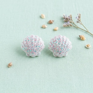 May include: A pair of seashell-shaped stud earrings featuring a white base covered in fine, multi-colored glitter in shades of pink, blue, and silver. The earrings have a textured, scalloped surface and a rounded, fan-like shell silhouette.