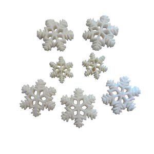May include: Seven white snowflake ornaments of varying sizes are arranged against a white background. The snowflakes have intricate, openwork designs, and a glittery texture, suggesting a festive or winter theme.
