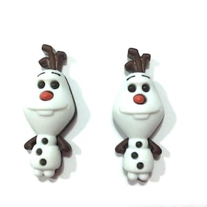 May include: Two white snowman-shaped charms with brown twig hair, black eyes, and a red nose. The snowman has three black buttons and brown arms. The charms are likely for jewelry or crafts.