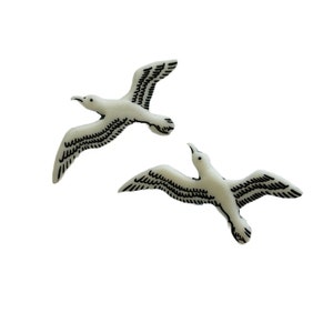 May include: Two off-white seagull figurines with black wing detailing are shown against a white background. The seagulls are in mid-flight, with their wings spread. The image is well-lit, highlighting the details of the birds.