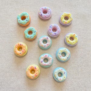 May include: A collection of twelve small, circular donut-shaped charms in various pastel colors including light blue, peach, lavender, yellow, and white. Each charm features a glossy finish and is decorated with tiny, multi-colored sprinkles.