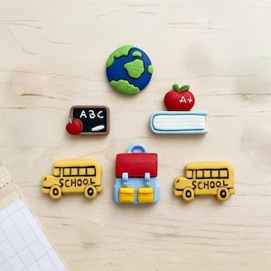 May include: A set of six school-themed decorative flat-back charms including a blue and green globe, a black chalkboard with ABC and an apple, a red apple on a blue book, a red and blue backpack, and two yellow school buses with black text.