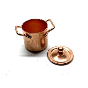 May include: A miniature copper cooking pot with two handles and a matching lid. The pot has a cylindrical shape and a shiny, metallic finish. The lid is round with a small knob on top. The items are likely for a dollhouse or miniature collection.