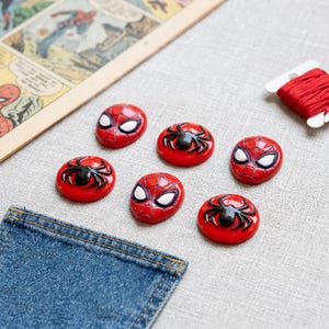 May include: A set of six circular buttons featuring red and black designs. Three buttons display a stylized black spider on a red background, while the other three buttons depict the red and black masked face of a comic book character.