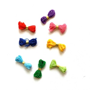 May include: A collection of colorful embroidery floss bows in red, purple, pink, yellow, blue, light blue, green, and light green. Each bow is secured with a gold-colored metal band, creating a decorative and vibrant display.