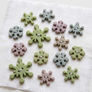 May include: A collection of snowflake-shaped buttons in various sizes and muted colors, including sage green, dusty rose, and slate gray. Each button features a central two-hole design with visible thread, arranged in a scattered pattern.