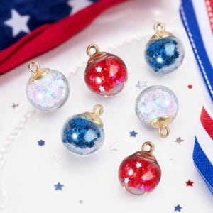 May include: Six spherical glass charms with gold-toned metal caps, each filled with star-shaped glitter in red, white, or blue. The charms are arranged in a cluster, featuring two red, two white, and two blue variations, all with small loops.