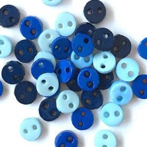 May include: A collection of small, round buttons in various shades of blue, including light blue, medium blue, and navy. Each button has two holes for sewing. The buttons are scattered on a white surface.