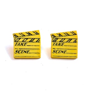 May include: A pair of yellow film clapperboard earrings. Each earring features the words "TAKE" and "SCENE" in black text. The clapperboards have a black and yellow striped design at the top.