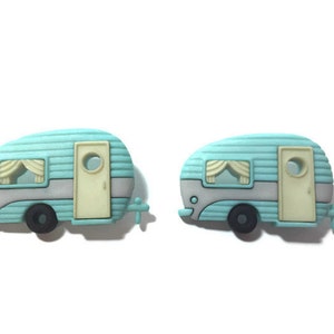 May include: Two turquoise and gray camper trailer charms. Each charm features a cream-colored door and window with a curtain. The wheels are black, and the trailer has a hitch. The charms are on a white background.