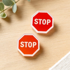 May include: Two octagonal pins featuring a red background with a white border and the word STOP printed in bold white capital letters. Each pin has a clear, raised acrylic-style edge, creating a distinct layered appearance on the surface.