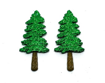 May include: Two glittery green Christmas tree-shaped embellishments with brown trunks. The trees have a layered design and are covered in sparkling green glitter. The background is plain white.