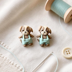 May include: Two identical buttons shaped like brown and white puppies wearing light blue overalls. The puppies have floppy ears, dark eyes, and small noses. The buttons are positioned near a wooden spool of light blue thread and a sewing needle.