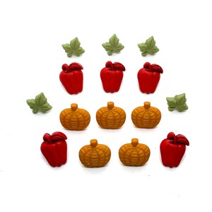 May include: A collection of decorative autumn-themed embellishments. The image features red apple-shaped and orange pumpkin-shaped buttons, along with green leaf-shaped buttons. These small, colorful buttons are arranged on a white background.