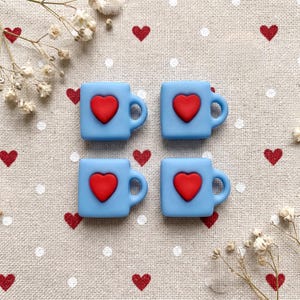 May include: Four square light blue mug-shaped charms, each featuring a raised red heart in the center. The charms are arranged in a two-by-two grid pattern, showcasing a smooth matte finish and a small handle on the right side of each piece.