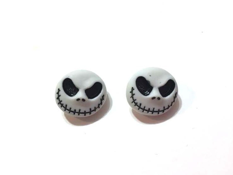 May include: A pair of white stud earrings featuring the face of Jack Skellington from The Nightmare Before Christmas. Each earring has black eyes and a stitched smile.