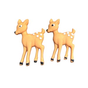 May include: Two small, tan-colored deer figurines with white spots and black hooves stand side-by-side. The deer have large eyes and are facing different directions. These decorative figurines are ideal for woodland-themed decor.