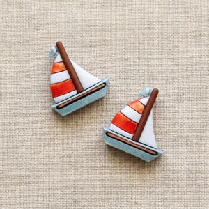 May include: Two sailboat-shaped buttons featuring a light blue hull, a brown mast, and sails with alternating horizontal stripes of white and bright orange. The buttons have a smooth, flat surface and are oriented in slightly different directions.