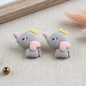 May include: Two gray elephant-shaped buttons, each featuring a small yellow bow on the head, a pink heart-shaped ear, and a raised trunk. The elephants have purple eyes and are positioned side by side on a textured fabric surface near a bead.