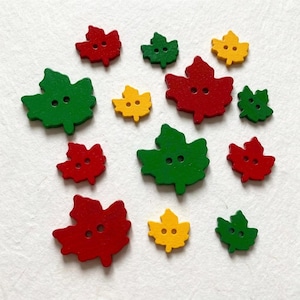 May include: A collection of flat, two-hole buttons shaped like maple leaves in various sizes and colors, including red, green, and yellow. The buttons are arranged in a scattered pattern, showcasing different leaf silhouettes and vibrant solid paint.